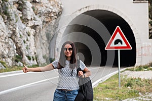 Young beautiful woman with black backpack hitchhiking standing near the road tunnel.