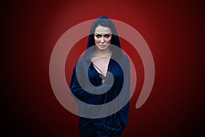 Young beautiful woman with a black hair and in the dark blue cloak with hood at the red background