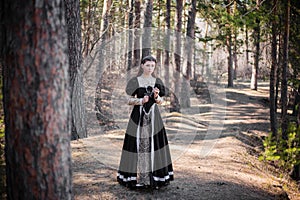 Young, beautiful woman in a black medieval dress with a steel rose in hand, walking through the forest. Attractive girl in a gothi