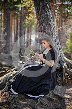 Young, beautiful woman in a black medieval dress with a steel rose in her hands, sitting in the woods on the roots of a tree. Attr