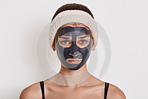 Young beautiful woman in black mud mask for face doing cosmetic procedures, looks sad, posing against white wall, wearing hair