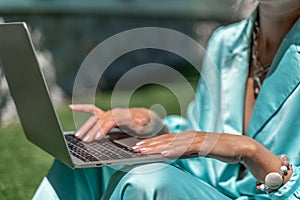 a young beautiful woman with blond curly hair in glasses and a blue dress sits on the grass in nature and uses a laptop,