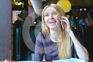 Young beautiful woman with blond hair talks on mobile phone and wide laugh, sitting at the table of cafe.
