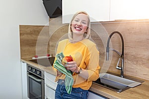 Young beautiful woman with blond red hair eating a juicy red apple while standing in her kitchen at home. Daily intake of vitamins