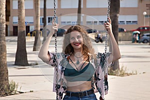 Young and beautiful woman, blonde, with curly hair and blue eyes, green top and jeans, sitting on a swing happy and flirtatious.