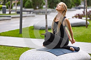 Young beautiful woman with blonde hair doing yoga exercise in green public park in summer morning, outdoors. Healthy