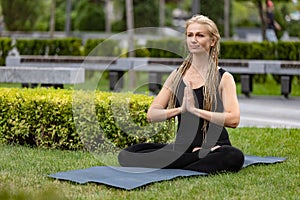 Young beautiful woman with blonde hair doing yoga exercise in green public park in summer morning, outdoors. Healthy