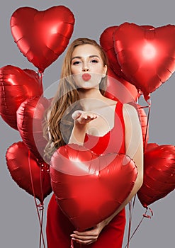 Young beautiful woman blowing kiss and holding red balloons. Model girl with red lips makeup wearing red dress on holiday party