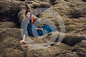 Young beautiful woman in blue dress sitting on the volcanic formations covered by green moss on a background green. Place for text