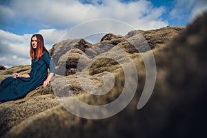 Young beautiful woman in blue dress sitting on the volcanic formations covered by green moss on a background sky. Place for text