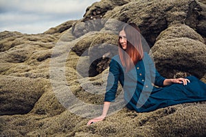 Young beautiful woman in blue dress sitting on the volcanic formations covered by green moss on a background sky. Place for text