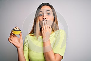 Young beautiful woman with blue eyes eating sweet chocolate cupcake over white background cover mouth with hand shocked with shame