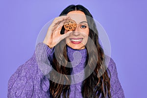 Young beautiful woman with blue eyes holding chocolate sweet coockie over eye with a happy face standing and smiling with a