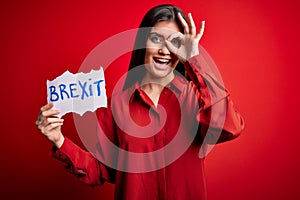 Young beautiful woman with blue eyes holding paper with brexit message over red background with happy face smiling doing ok sign