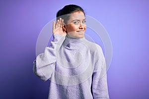 Young beautiful woman with blue eyes wearing casual turtleneck sweater over pink background smiling with hand over ear listening