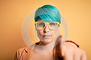 Young beautiful woman with blue fashion hair wearing casual glasses over yellow background pointing with finger to the camera and