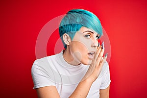 Young beautiful woman with blue fashion hair wearing casual t-shirt over red background hand on mouth telling secret rumor,