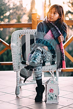 Young beautiful woman in a blue sweater with colorful scarf sitting on a white chair during cold weather outside the balcony