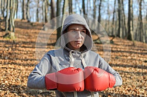 Young beautiful woman boxer in a tracksuit with a hood on his head in red boxing gloves trains in the forest in nature