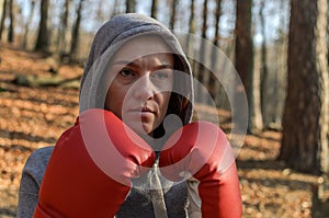Young beautiful woman boxer in a tracksuit with a hood on his head in red boxing gloves trains in the forest in nature