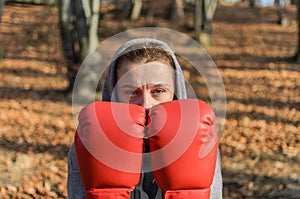 Young beautiful woman boxer in a tracksuit with a hood on his head in red boxing gloves trains in the forest in nature