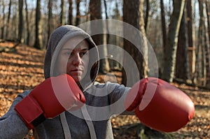 Young beautiful woman boxer in a tracksuit with a hood on his head in red boxing gloves trains in the forest in nature