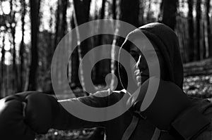 Young beautiful woman boxer in a tracksuit with a hood on his head in red boxing gloves trains in the forest in nature