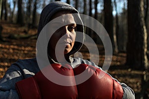 Young beautiful woman boxer in a tracksuit with a hood on his head in red boxing gloves trains in the forest in nature