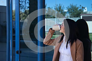 Young beautiful woman in brown coat drinking coffee, leaning against her office door at breakfast time. Business concept, working