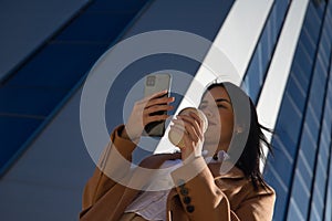 Young, beautiful woman in brown coat and glass of coffee in hand, checking social networks on her cell phone on her way out of the