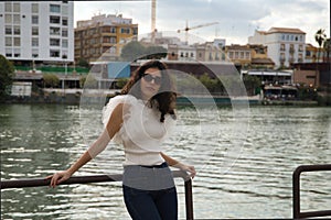 Young and beautiful woman, brunette, with curly hair, wearing white shirt, jeans and sunglasses, leaning on a railing by the river