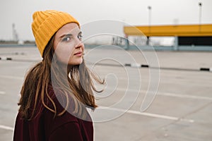 Young beautiful woman in burgundy coat and yellow hat walks along the roof of the parking lot. Loneliness, mindfulness.