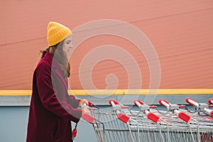 Young beautiful woman in a burgundy coat and yellow hat walks on the roof of supermarket parking lot and takes trolley.