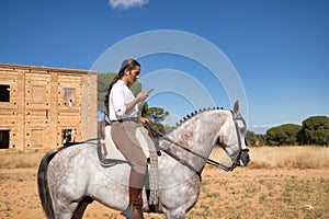Young beautiful woman checking social networks on her cell phone, riding her horse in the countryside. Concept horse riding,