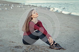 Young beautiful woman with closed eyes, long hair, in black jeans and red shirt, sitting on sand on beach among seagulls birds