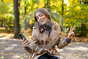 Young beautiful woman in a coat writes a message in the phone while sitting on a bench in the autumn park