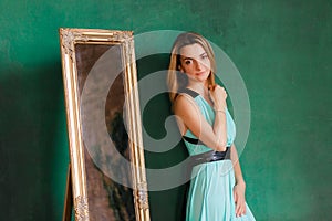 Young beautiful woman in cocktail dress standing near vintage mirror. With green empty wall on background, copyspace