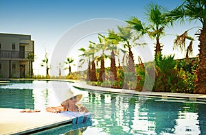 Young beautiful woman with cocktail glass, hat, bikini in blue water swimming pool at sunset party