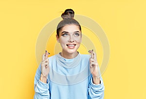 Young beautiful woman crossing her fingers and wishing for good luck over yellow background.