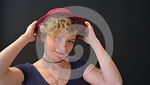 Young beautiful woman with curly blonde hair touching her hat and smiling at camera, isolated on black background