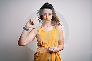 Young beautiful woman with curly hair controlling weight using tape mesure with angry face, negative sign showing dislike with