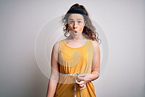 Young beautiful woman with curly hair controlling weight using tape mesure scared in shock with a surprise face, afraid and