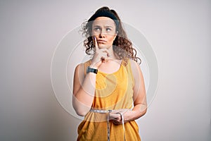 Young beautiful woman with curly hair controlling weight using tape mesure serious face thinking about question, very confused