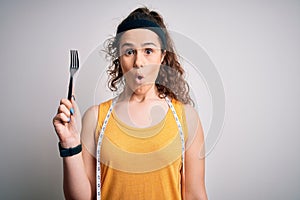 Young beautiful woman with curly hair doing diet holding fork and tape measure scared in shock with a surprise face, afraid and