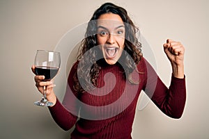 Young beautiful woman with curly hair drinking glass of red wine over white background screaming proud and celebrating victory and