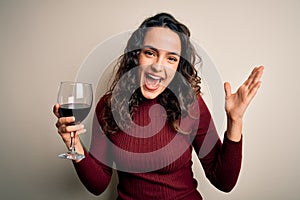 Young beautiful woman with curly hair drinking glass of red wine over white background very happy and excited, winner expression