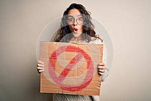 Young beautiful woman with curly hair holding banner with prohibited signal message scared in shock with a surprise face, afraid