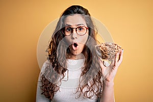 Young beautiful woman with curly hair holding bowl with healthy corn flakes cereals scared in shock with a surprise face, afraid