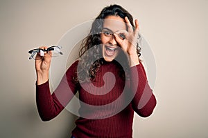 Young beautiful woman with curly hair holding glasses over isolated white background with happy face smiling doing ok sign with