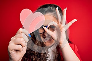 Young beautiful woman with curly hair holding paper heart over isolated red background with happy face smiling doing ok sign with